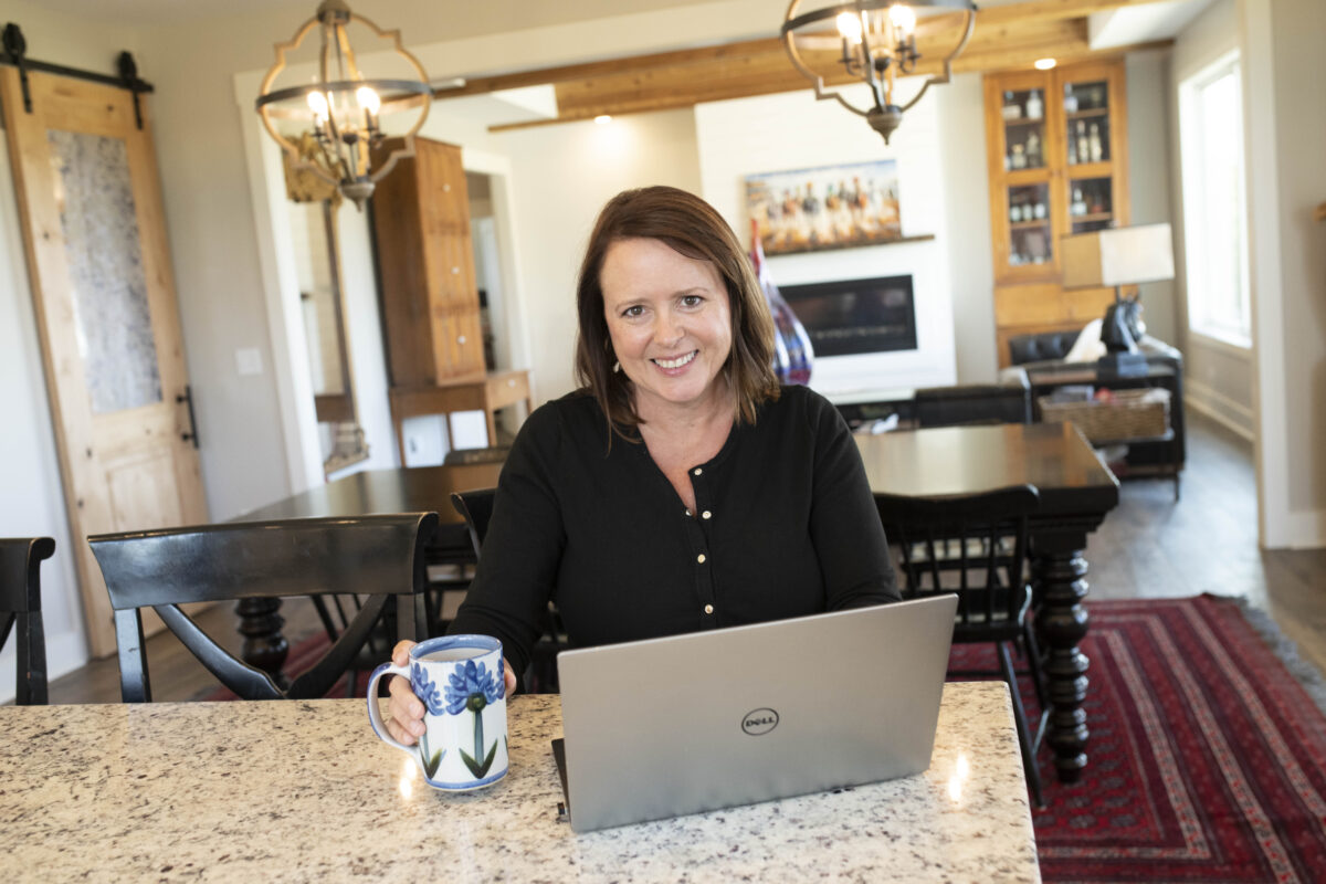 Smiling attractive woman sitting at kitchen island with computer and coffee cup with beautiful living room in the background